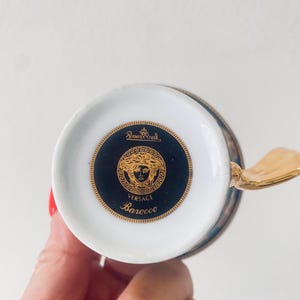 May include: A white and gold teacup with a gold handle. The bottom of the cup features a black and gold medallion with the Versace logo and the word "Barocco". The cup is part of the Rosenthal Versace collection.