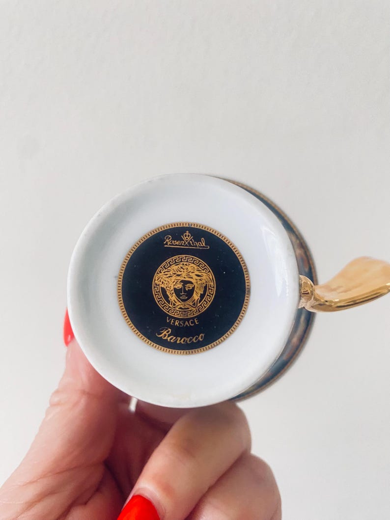 May include: A white and gold teacup with a gold handle. The bottom of the cup features a black and gold medallion with the Versace logo and the word "Barocco". The cup is part of the Rosenthal Versace collection.