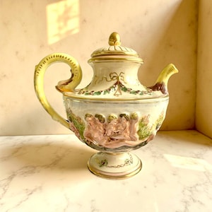 May include: An antique, ornate teapot with a cream-colored base and lid, accented with gold trim. The teapot features a curved yellow handle and spout. The body of the teapot is decorated with a relief of cherubs and floral designs. The teapot sits on a pedestal base.