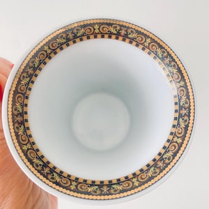 May include: White teacup with a decorative border featuring gold, black, and brown ornate scrollwork. The cup's rim has a detailed pattern, and the interior is plain white. The design suggests a vintage or luxury aesthetic.
