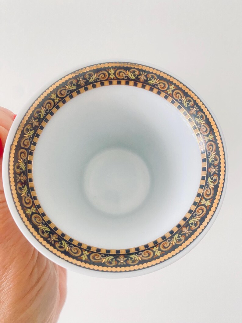 May include: White teacup with a decorative border featuring gold, black, and brown ornate scrollwork. The cup's rim has a detailed pattern, and the interior is plain white. The design suggests a vintage or luxury aesthetic.