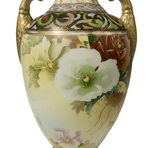 May include: A decorative porcelain vase with a cream-colored background and floral designs. The vase features a large white flower, red and pink roses, and gold-colored handles and trim. The vase has a bulbous shape.