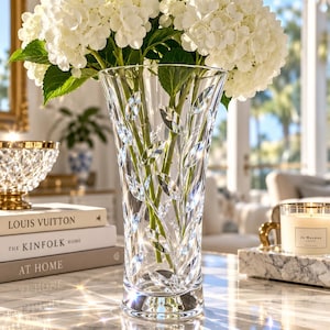 May include: A clear glass vase featuring a cut leaf pattern, filled with a large bouquet of white hydrangea flowers. The vase has a flared rim and a thick base, reflecting light to create bright starburst patterns on the marble surface below.