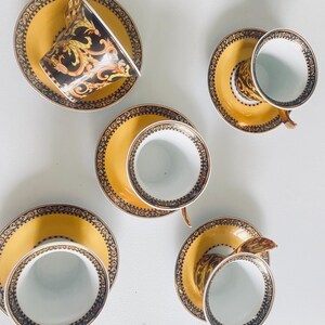 May include: A set of six teacups and saucers. The saucers are yellow with a decorative black and gold border. The teacups are white inside with a black and gold design on the outside. One cup is tilted, showing the interior design.