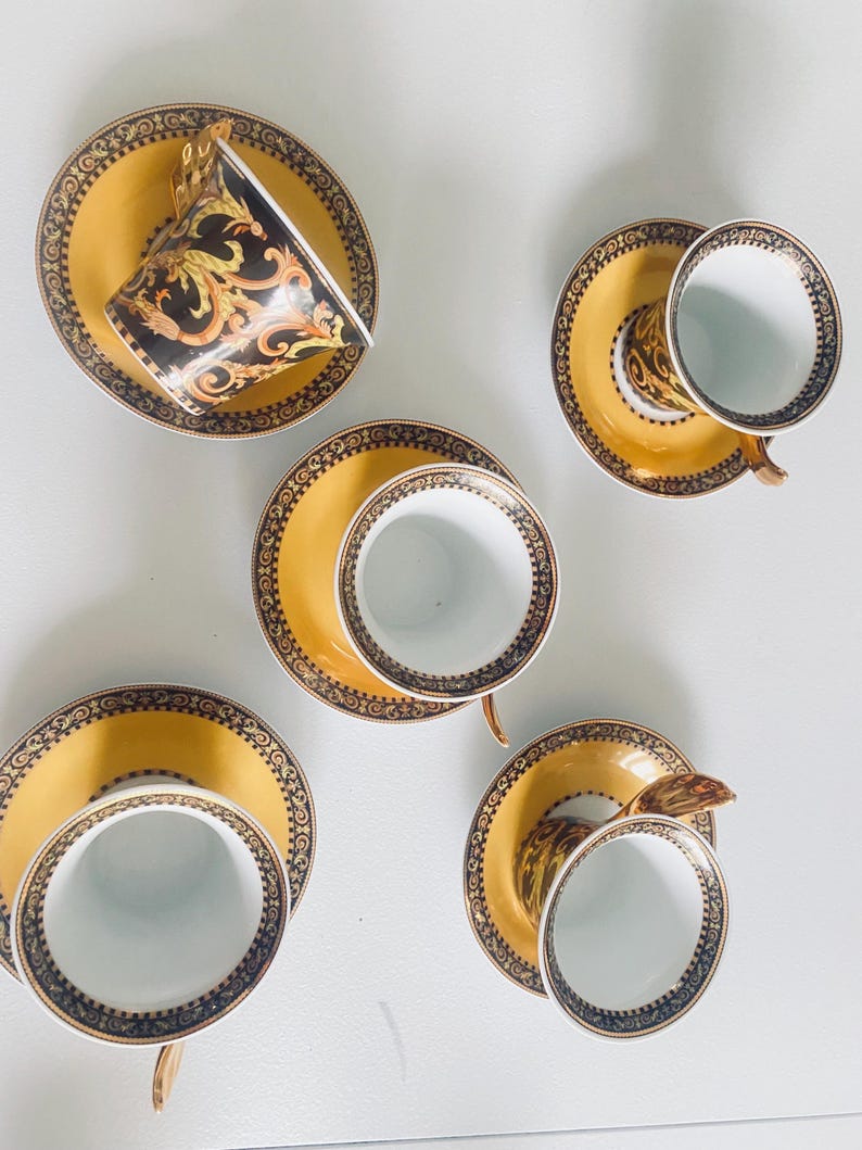 May include: A set of six teacups and saucers. The saucers are yellow with a decorative black and gold border. The teacups are white inside with a black and gold design on the outside. One cup is tilted, showing the interior design.