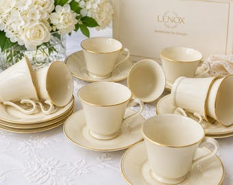 May include: A set of ivory-colored teacups and saucers with gold trim. The cups are arranged on a white tablecloth, with a vase of white flowers in the background. The Lenox box is visible.
