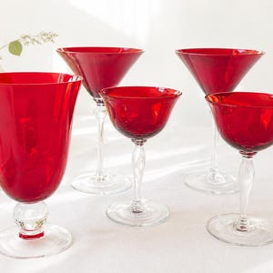 May include: A collection of five red glass drinking glasses. The glasses vary in shape, including a tall tumbler, martini glasses, and stemmed glasses. The glasses are arranged on a white surface, with a vase of flowers in the background.