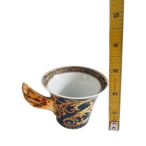 May include: A decorative teacup with a gold handle and a dark blue body. The cup features a gold and yellow ornate design with a black and gold rim. The cup is approximately 3 inches tall.