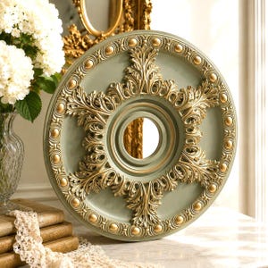 May include: A circular ceiling medallion featuring a sage green center and an ornate gold leaf finish. The design includes a central circular opening surrounded by intricate acanthus leaf carvings and a border of raised gold spheres and detailing.