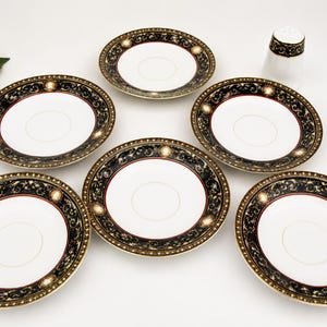 May include: A set of six white porcelain saucers with a black, gold, and red decorative border. A matching salt shaker with a similar design is also present. The saucers are arranged on a white surface.
