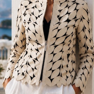 May include: Cream-colored jacket with a black leaf-like pattern and a scalloped hem. The jacket has a zipper closure and is paired with white pants. The model is wearing sunglasses and pearl earrings.