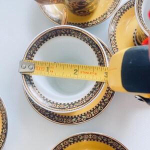May include: A set of ornate teacups and saucers. The teacups are white inside with a gold handle and a decorative gold and black pattern around the rim. The saucers are yellow with a matching pattern. The cup's diameter is approximately 3 inches.