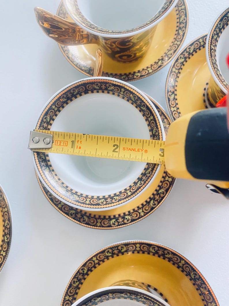 May include: A set of ornate teacups and saucers. The teacups are white inside with a gold handle and a decorative gold and black pattern around the rim. The saucers are yellow with a matching pattern. The cup's diameter is approximately 3 inches.