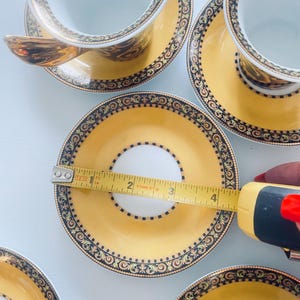May include: A set of yellow and gold teacups and saucers with ornate black and gold detailing. The saucer measures approximately 4 inches in diameter. The teacups have a white interior and gold handles. The set is on a white surface.