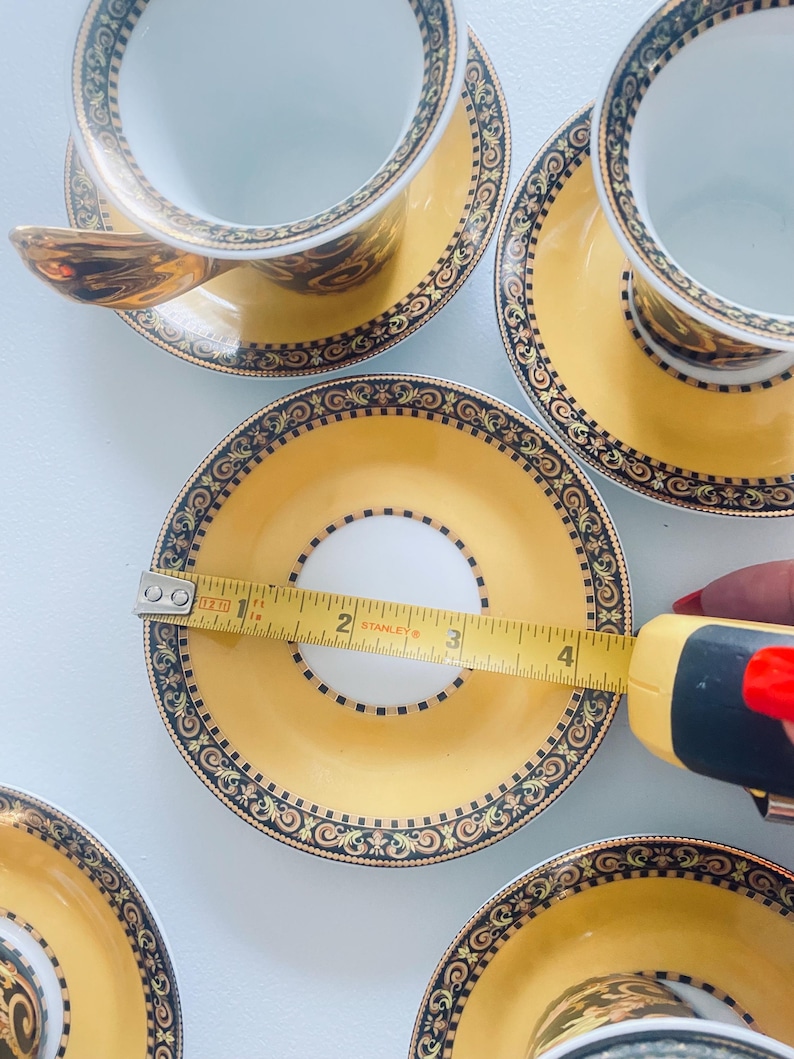 May include: A set of yellow and gold teacups and saucers with ornate black and gold detailing. The saucer measures approximately 4 inches in diameter. The teacups have a white interior and gold handles. The set is on a white surface.