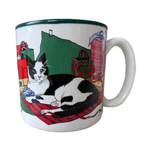 May include: White ceramic mug featuring a black and white cat lying on a red sled. The mug is decorated with Christmas gifts and a green border. The handle is white.