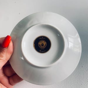 May include: White porcelain saucer with a gold and black Versace logo on the underside. The logo features a circular design with the brand name and a decorative emblem. The saucer is round and has a slightly raised edge.