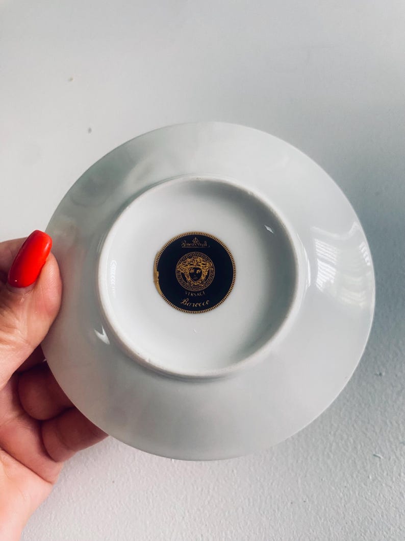May include: White porcelain saucer with a gold and black Versace logo on the underside. The logo features a circular design with the brand name and a decorative emblem. The saucer is round and has a slightly raised edge.