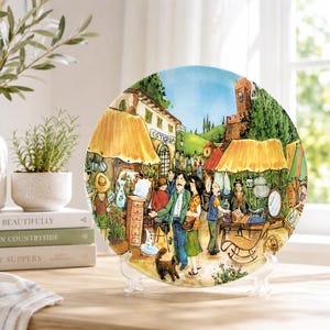 May include: A round decorative ceramic plate featuring a colorful illustration of a bustling outdoor market scene with people, yellow awnings, a stone tower, and a building labeled Osteria. The artwork uses vibrant tones of yellow, green, and blue.
