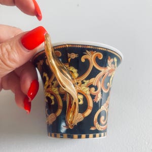 May include: A black and gold teacup with a gold handle. The teacup features an ornate design with gold and tan accents. The handle is a curved, gold design. The teacup is held by a person with red nail polish.