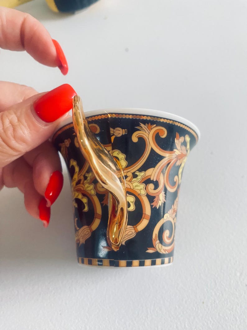 May include: A black and gold teacup with a gold handle. The teacup features an ornate design with gold and tan accents. The handle is a curved, gold design. The teacup is held by a person with red nail polish.