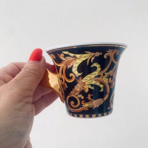 May include: A black and gold teacup with ornate floral and scrollwork designs. The cup has a gold handle and a white interior rim. The design features yellow and gold accents. The cup is held against a white background.