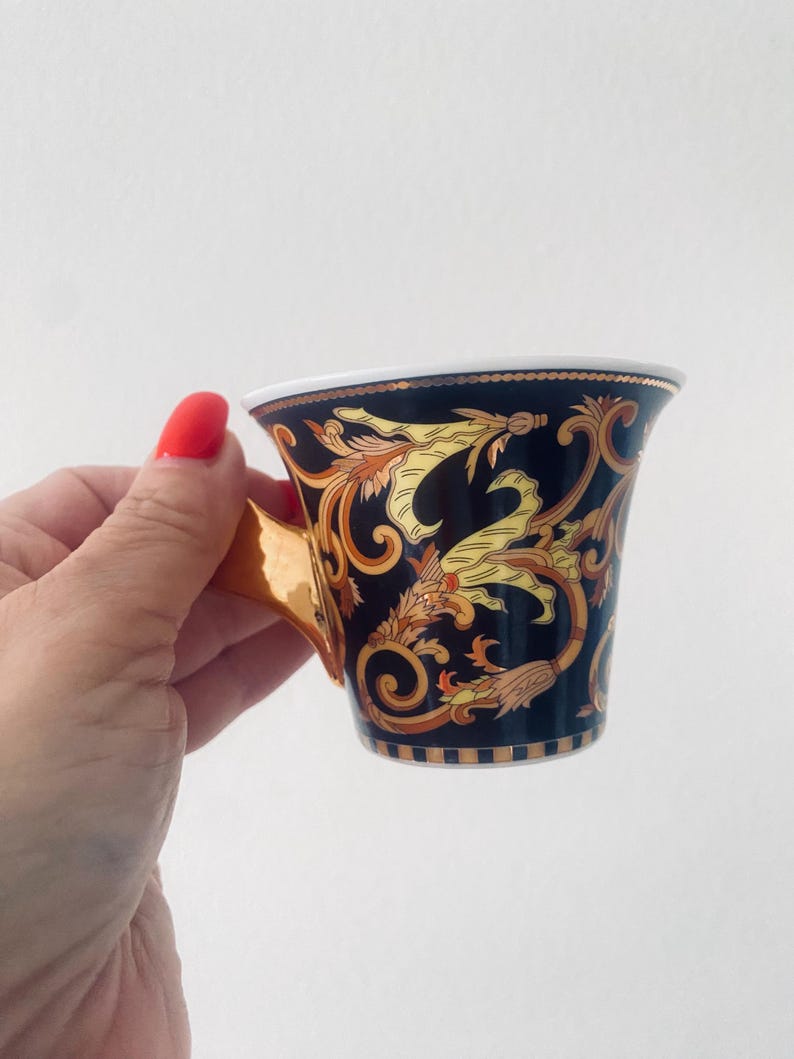 May include: A black and gold teacup with ornate floral and scrollwork designs. The cup has a gold handle and a white interior rim. The design features yellow and gold accents. The cup is held against a white background.