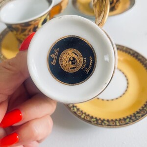 May include: A white teacup with a gold handle and a black and gold design. The bottom of the cup features a black circle with a gold emblem and the words "Versace Barocco". The cup is part of a set with a matching saucer.