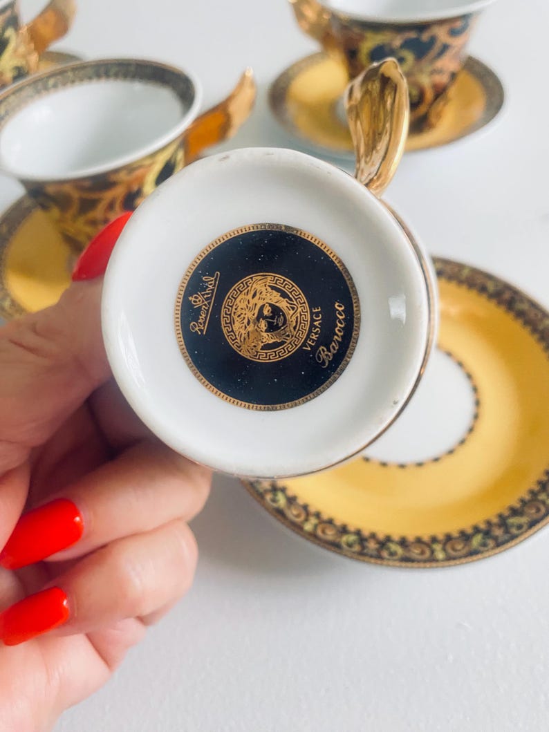 May include: A white teacup with a gold handle and a black and gold design. The bottom of the cup features a black circle with a gold emblem and the words "Versace Barocco". The cup is part of a set with a matching saucer.