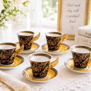 May include: A set of six decorative teacups and saucers. The cups are black with gold scrollwork and wing-shaped handles. The saucers are yellow with a black and gold border. A gold-framed sign reads "Start each day with a grateful heart."