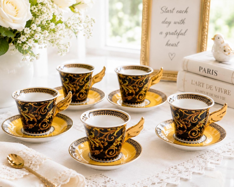 May include: A set of six decorative teacups and saucers. The cups are black with gold scrollwork and wing-shaped handles. The saucers are yellow with a black and gold border. A gold-framed sign reads "Start each day with a grateful heart."
