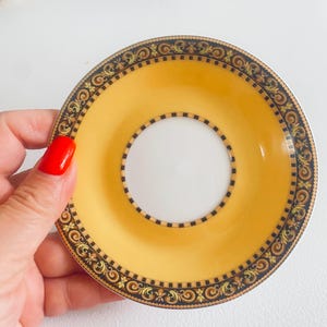 May include: A yellow and white saucer with an ornate black and gold border. The saucer has a central white circle and a wide yellow band. The rim features a repeating scroll and geometric pattern. A decorative piece for serving tea or coffee.