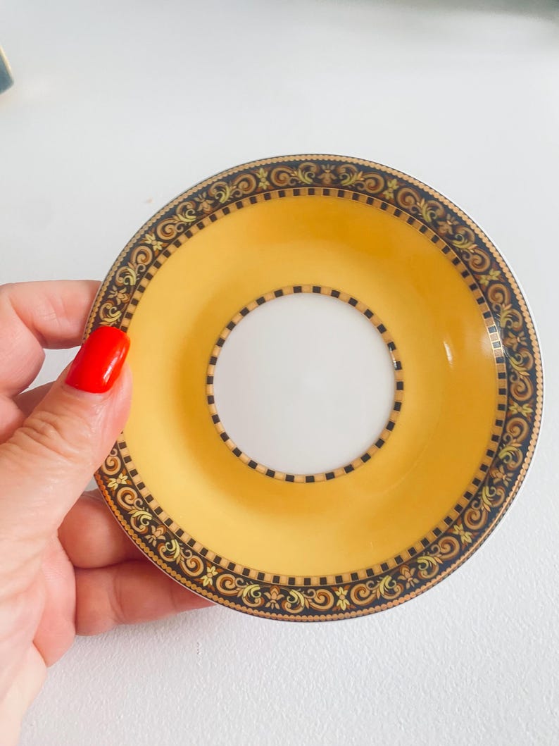 May include: A yellow and white saucer with an ornate black and gold border. The saucer has a central white circle and a wide yellow band. The rim features a repeating scroll and geometric pattern. A decorative piece for serving tea or coffee.