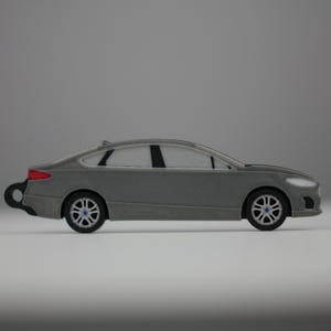 May include: A gray toy car with black wheels and blue accents. The car has a sleek design with a red taillight and a wind-up key on the back. The car is a miniature replica of a modern sedan.