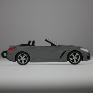 Puede incluir: Modelo de coche descapotable gris con ruedas negras e interior negro. El coche tiene un diseño elegante con un perfil bajo y un frontal detallado. El piloto trasero es rojo.