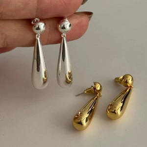 Puede incluir: Dos pares de pendientes de lágrima. Un par es plateado, con una pequeña bola en la parte superior y una forma de lágrima larga y lisa. El otro par es dorado, con un diseño similar. Los pendientes se muestran sobre una superficie blanca.