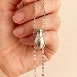 May include: A silver-toned necklace featuring a small, smooth, amphora-shaped pendant suspended from a delicate link chain. The pendant has two small handles on the sides and a polished, reflective surface that catches the light.