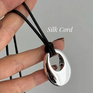 May include: A silver-toned pendant with a smooth, polished, oval-shaped design featuring a central cutout. The pendant is suspended from a black cord, with the text Silk Cord visible in white lettering next to the jewelry piece.