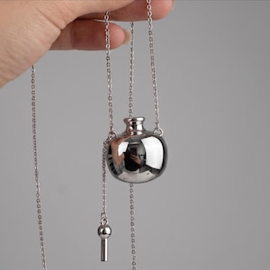 Puede incluir: Un collar con colgante de metal plateado que presenta un pequeño dije en forma de botella esférica con acabado de espejo, suspendido de una cadena fina. El diseño incluye un elemento colgante secundario con una cuenta cilíndrica al final.