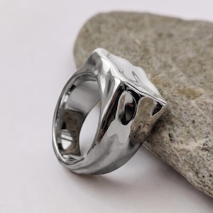 May include: A polished silver-tone ring with an irregular, melted-look surface. The band features a thick, organic shape with deep indentations and smooth, reflective contours that create a liquid metal appearance across the entire outer surface.
