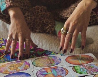 May include: A spread of tarot cards with colorful illustrations on a patterned surface. The cards are oval-shaped and feature images of a honeycomb, a forest, a mushroom, a dragonfly, flowers, and a tree.