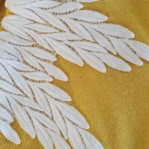 Puede incluir: Primer plano de una tela amarillo mostaza con detalles blancos bordados en forma de alas. Las alas est&aacute;n hechas de elementos delicados en forma de hojas superpuestas, creando un efecto texturizado y tridimensional. El dise&ntilde;o sugiere un proyecto artesanal o textil.