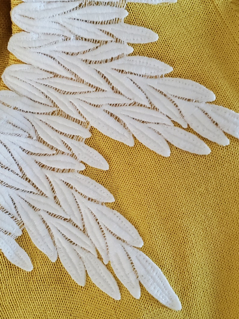 Puede incluir: Primer plano de una tela amarillo mostaza con detalles blancos bordados en forma de alas. Las alas est&aacute;n hechas de elementos delicados en forma de hojas superpuestas, creando un efecto texturizado y tridimensional. El dise&ntilde;o sugiere un proyecto artesanal o textil.