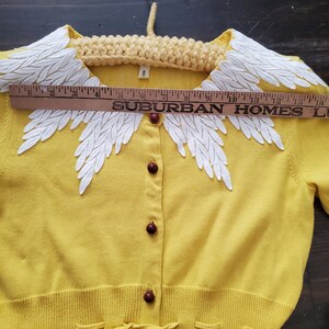 Puede incluir: C&aacute;rdigan amarillo vintage con cuello de encaje blanco con forma de hojas. El c&aacute;rdigan tiene botones marrones y un lazo en la cintura. Una regla muestra el texto "SUBURBAN HOMES". La etiqueta de talla indica "8".