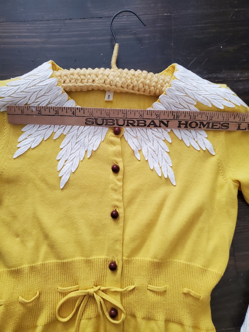 Puede incluir: C&aacute;rdigan amarillo vintage con cuello de encaje blanco con forma de hojas. El c&aacute;rdigan tiene botones marrones y un lazo en la cintura. Una regla muestra el texto "SUBURBAN HOMES". La etiqueta de talla indica "8".