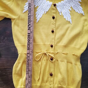 Puede incluir: C&aacute;rdigan amarillo con un dise&ntilde;o blanco en forma de hojas alrededor del cuello. Tiene botones marrones oscuros y un lazo en la cintura. Una regla de madera se coloca junto al c&aacute;rdigan, midiendo aproximadamente 56 cm.