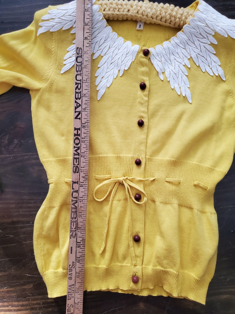 Puede incluir: C&aacute;rdigan amarillo con un dise&ntilde;o blanco en forma de hojas alrededor del cuello. Tiene botones marrones oscuros y un lazo en la cintura. Una regla de madera se coloca junto al c&aacute;rdigan, midiendo aproximadamente 56 cm.