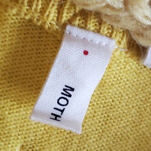 Puede incluir: Primer plano de una etiqueta de tela blanca con la palabra "MOTH" impresa en negro. Un peque&ntilde;o punto rojo est&aacute; encima del texto. La etiqueta est&aacute; cosida a una tela de punto amarilla, con una parte de un art&iacute;culo de punto crema visible.