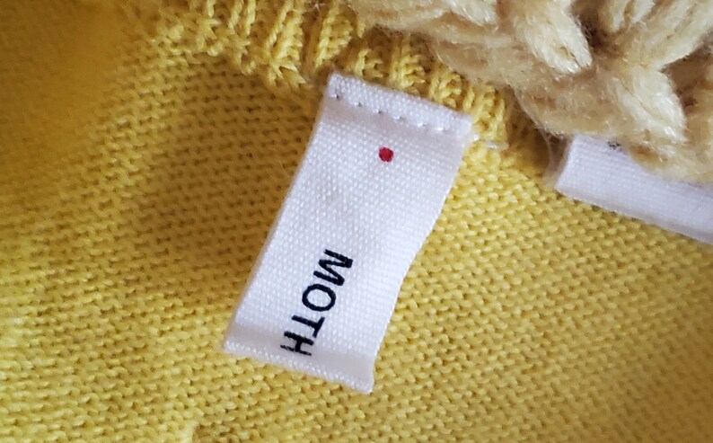 Puede incluir: Primer plano de una etiqueta de tela blanca con la palabra "MOTH" impresa en negro. Un peque&ntilde;o punto rojo est&aacute; encima del texto. La etiqueta est&aacute; cosida a una tela de punto amarilla, con una parte de un art&iacute;culo de punto crema visible.