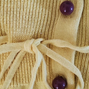 Puede incluir: Primer plano de un c&aacute;rdigan de punto amarillo p&aacute;lido. El c&aacute;rdigan tiene un cierre de lazo y dos botones de color p&uacute;rpura oscuro. El tejido de punto tiene una textura acanalada. La corbata est&aacute; hecha del mismo material que el c&aacute;rdigan.